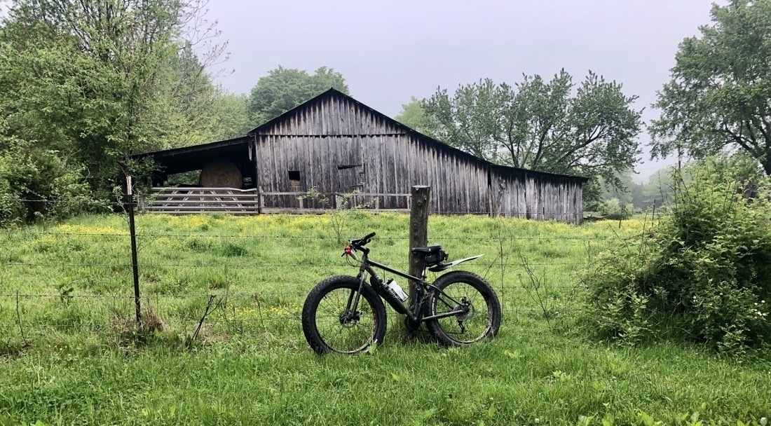 A black bike with oversized tires leans against a fencepost, an old barn in the background