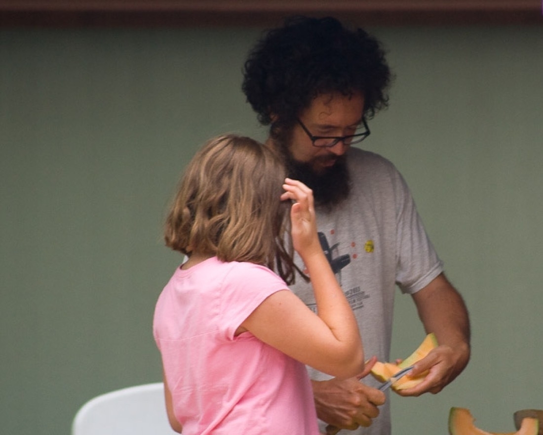 A beardy man with glasses and large fuzzy black hair is cutting a cantaloupe. A girl with back to camera is standing next to him