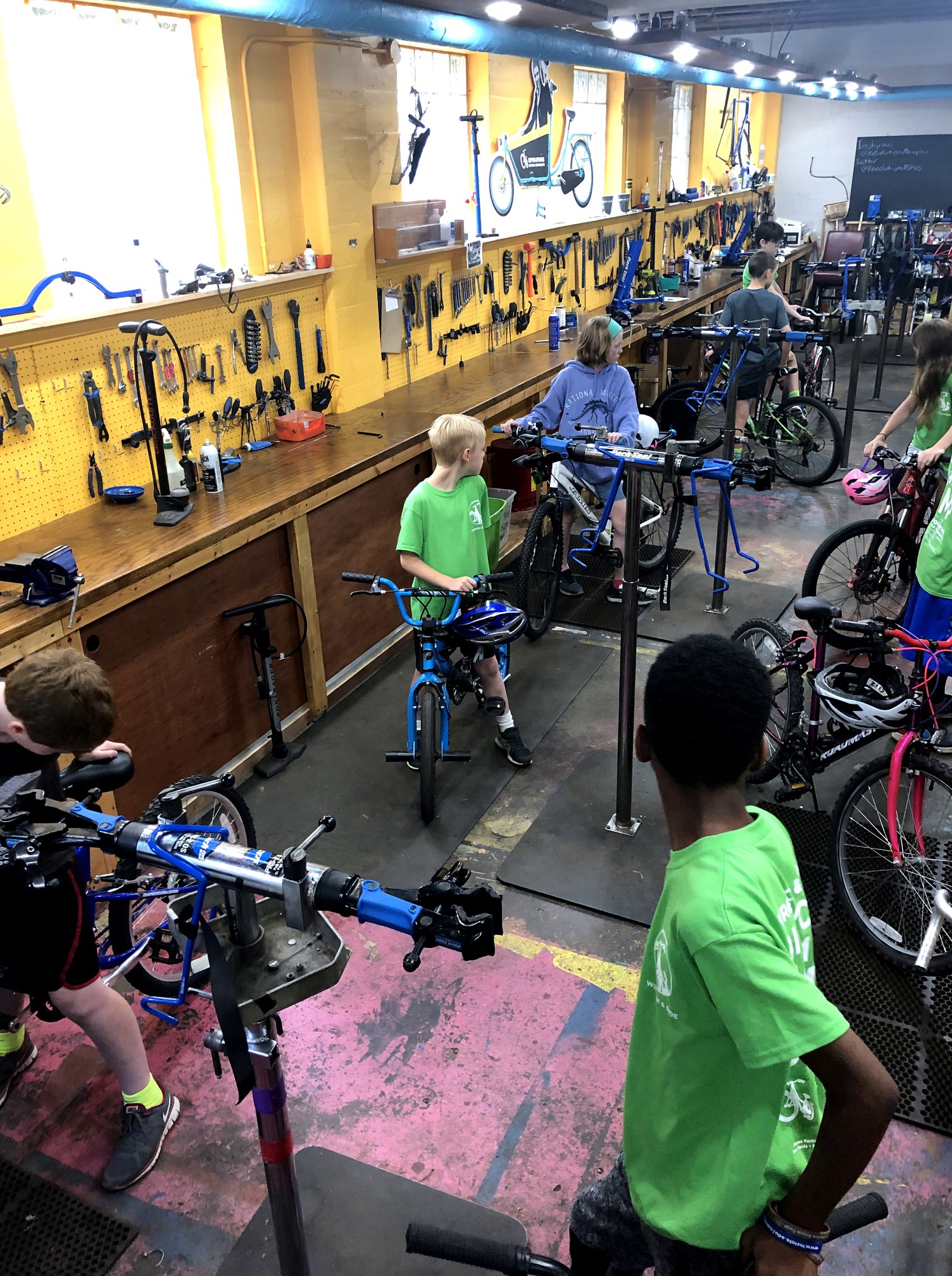 Kids at a bicycle repair workshop are repairing bikes in with shelves of bike tools behind them