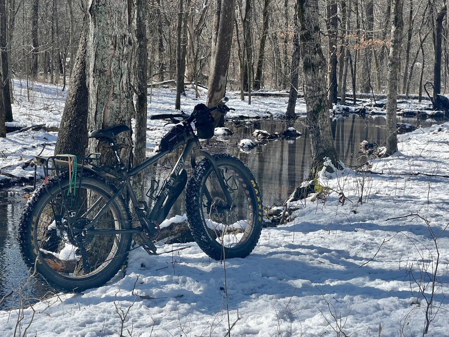 A fat tire bike leans against a tree on a snow covered trail in the woods. A creek is behind the bike.