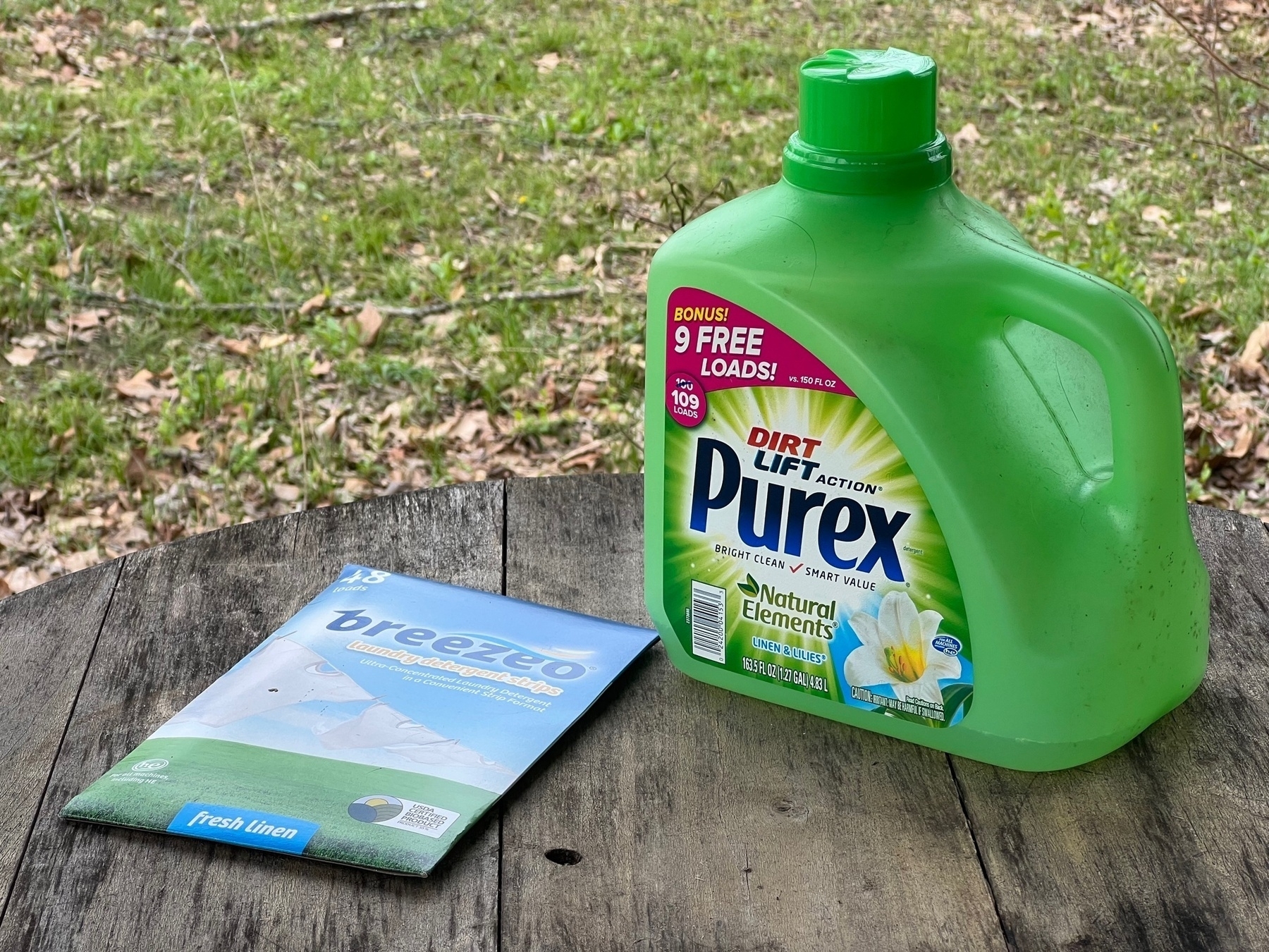a paper board container of laundry detergent strips is in slim package sitting on the table next to a green plastic jug of liquid laundry detergent.