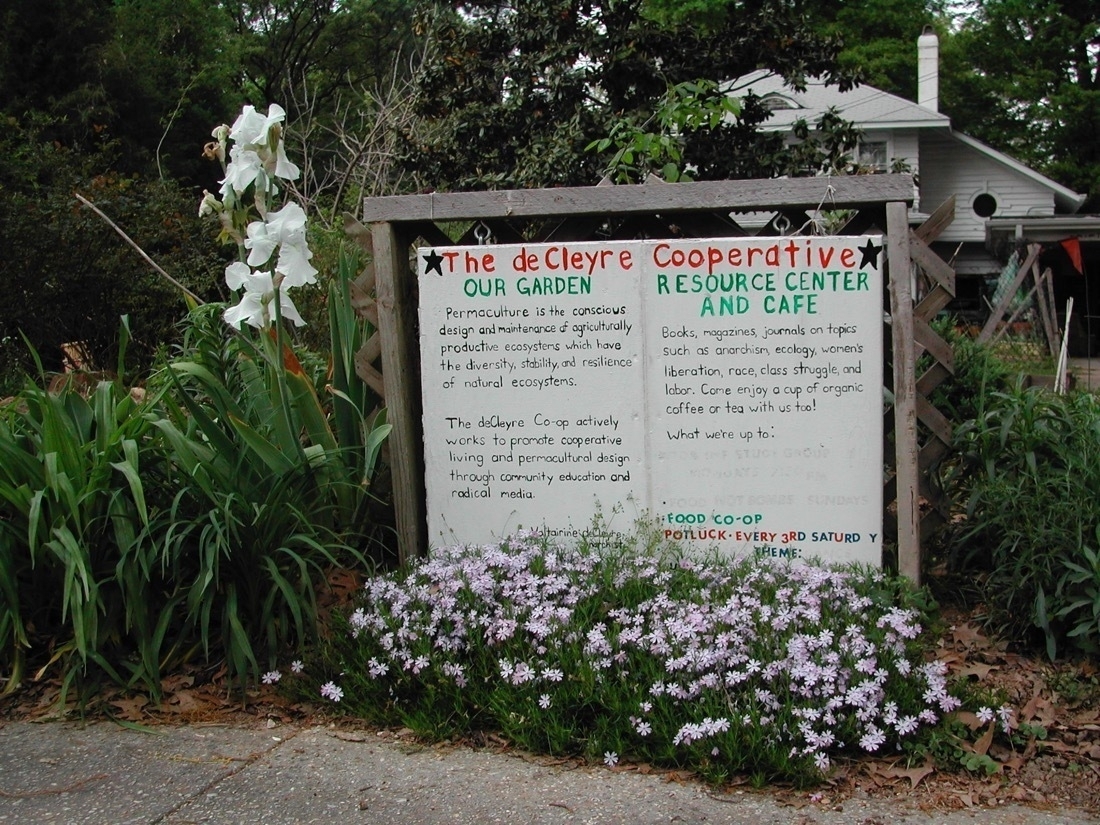A handpainted white sign in front of a house with this wording: The deCleyre Cooperative OUR GARDEN RESOURCE CENTER AND CAFE Permaculture is the conscious design and maintenance of agriculturally productive ecosystems which have the diversity, stability, and resilience of natural ecosystems. Books, magazines, journals on topicssuch as anarchism, ecology, women's liberation, race, class struggle, and labor. Come enjoy a cup of organic coffee or tea with us too! The deCleyre Co-op actively works to promote cooperative living and permacultural design through community education and radical media.